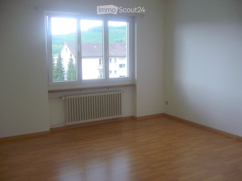 Empty room with wooden flooring, white walls, large windows, radiator, and a view of a house and landscape.