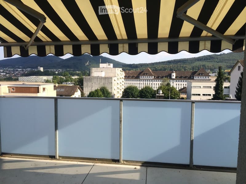 Balcony with white panels, metal railing, and a striped awning, overlooking a cityscape with buildings and mountains.