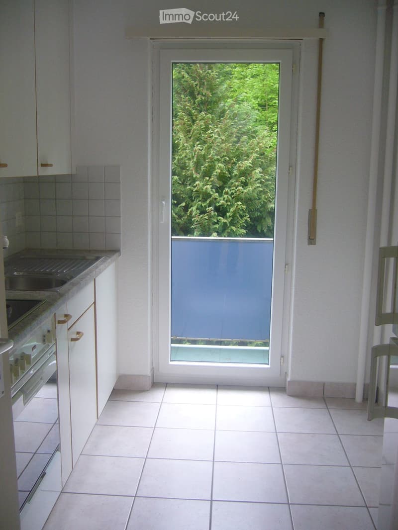 Modern kitchen with white tiled floor, white cabinets, and a large sliding glass door leading to a balcony with a view of greenery.