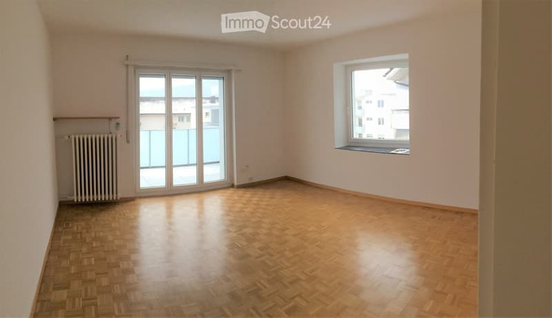 Empty room with wooden flooring, large sliding glass door leading to a balcony, and a window with a view of neighboring buildings.