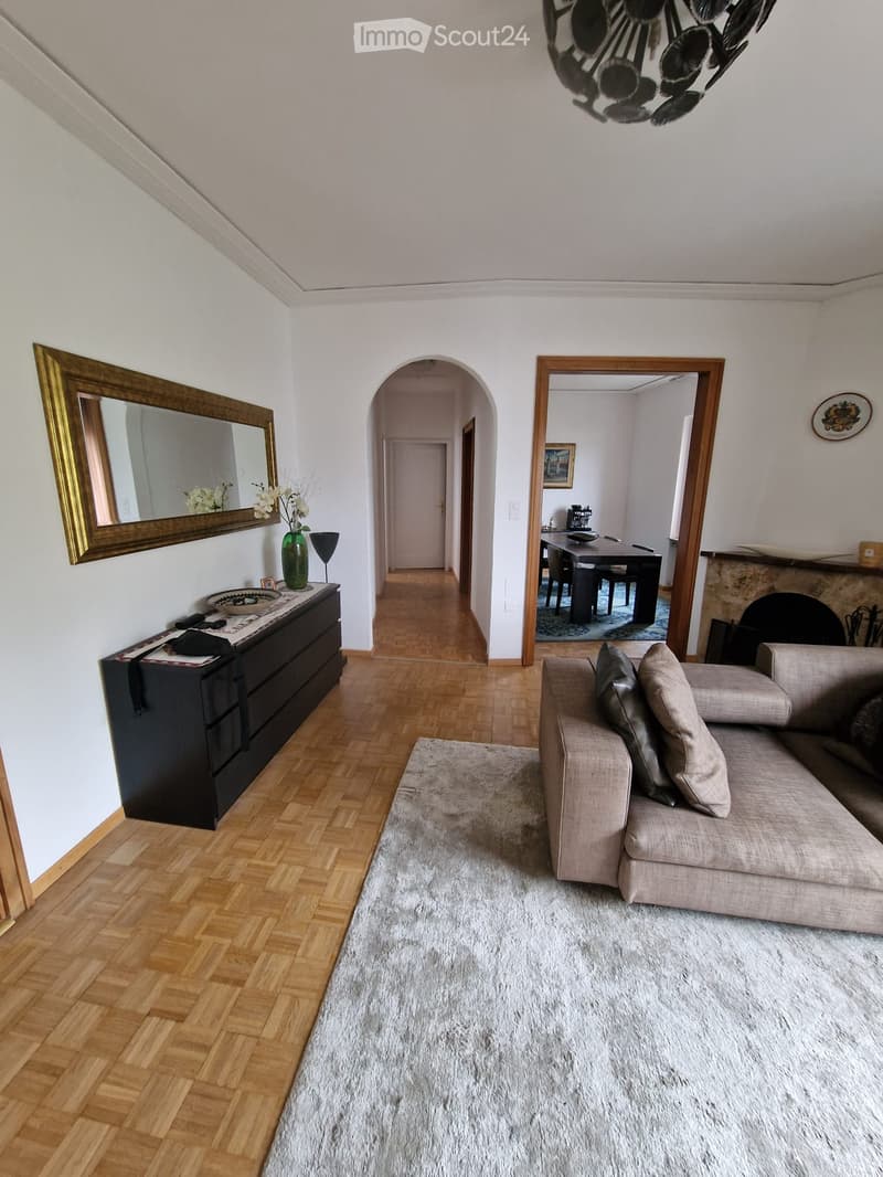 Living room with a beige sofa, coffee table, large mirror, vase with flowers, and a view into a dining area with a table and chairs.