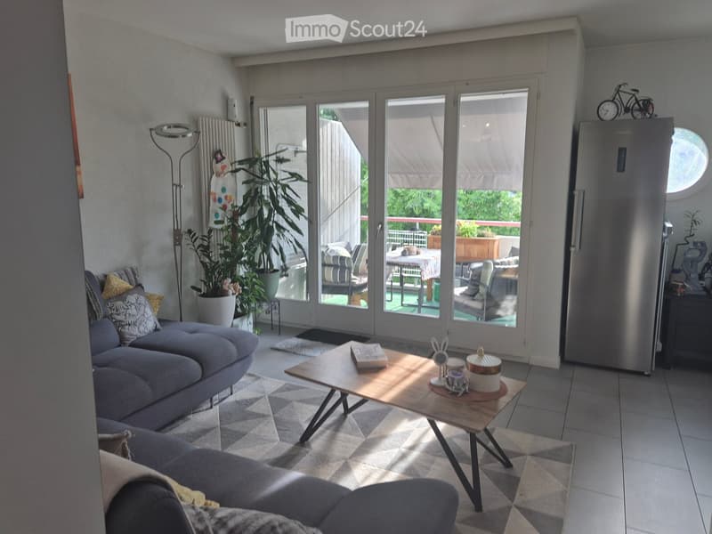 Bright living room with large sliding glass doors leading to a balcony, grey sectional sofa, wooden coffee table, floor lamp, and a refrigerator visible in the background.