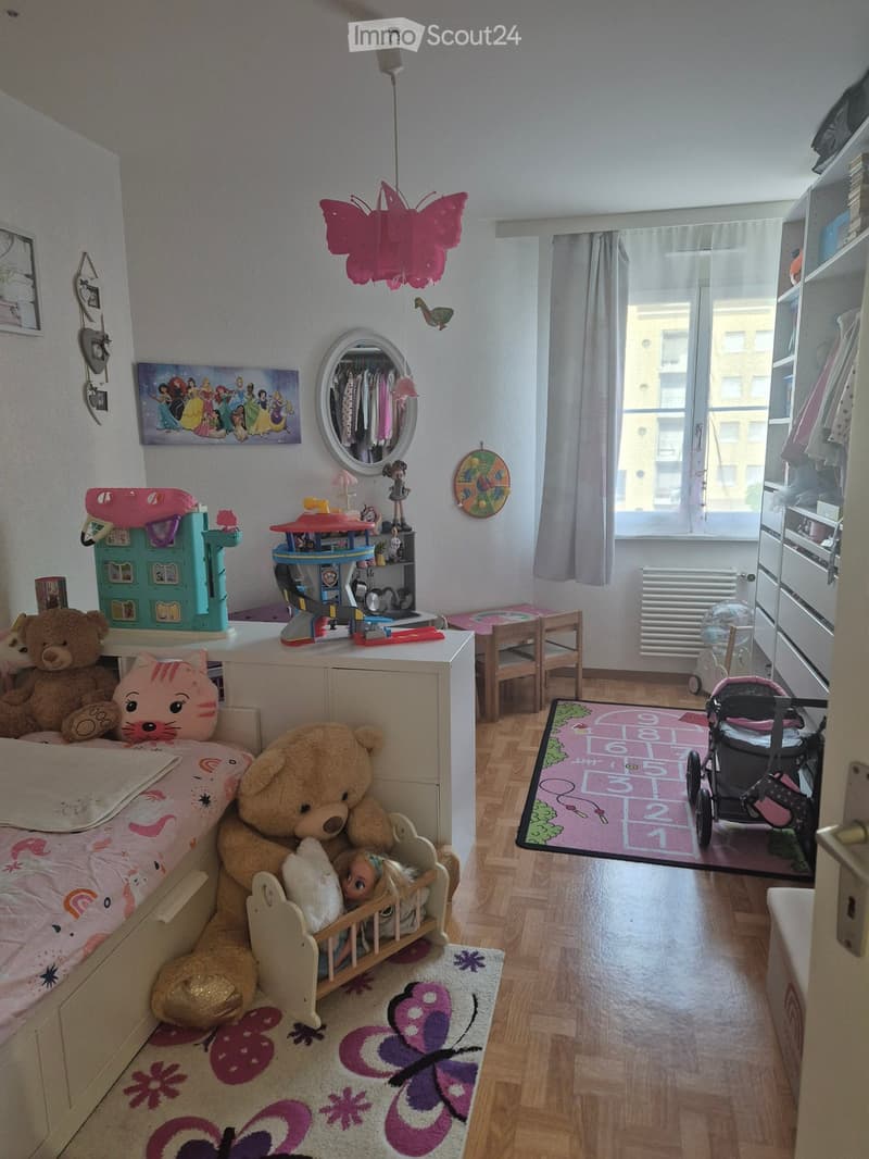 A well-lit children's bedroom with a bed, teddy bears, a doll, a toy house, a mirror, a wardrobe, a chair, a table, and various toys. The room features a large window with curtains, a radiator, and a baby stroller near the door.