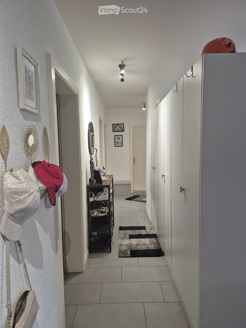 A well-lit hallway with tiled flooring, white walls, and several doors leading to different rooms. There are decorative items on the walls, a small shelf with shoes, and a small rug on the floor.