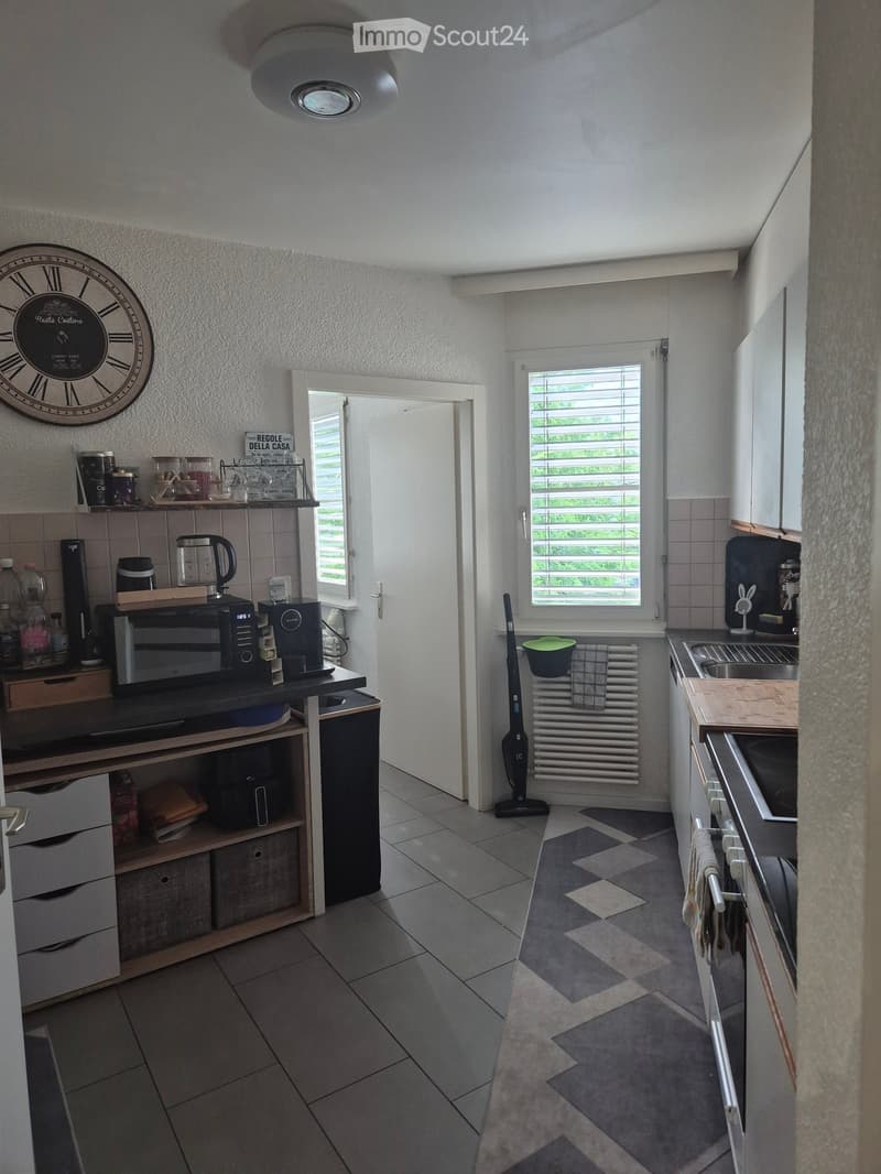 Modern kitchen with grey tiled floor, white walls, and fitted cabinets. Includes microwave, coffee maker, fridge, oven, sink, and various kitchen items on the counter.
