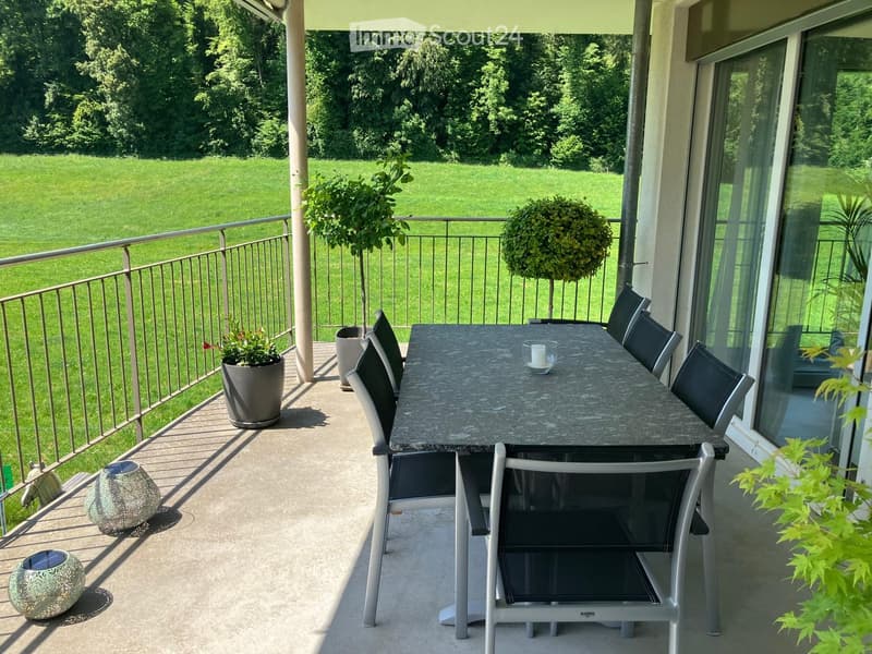 Balcony with a dining table and chairs, surrounded by potted plants and a view of a green lawn.