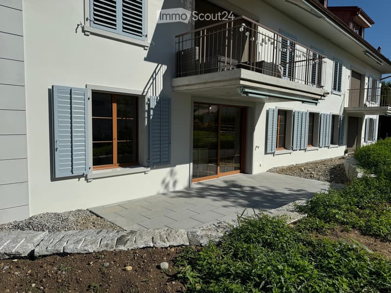Two-story house with light gray shutters, large windows, and a balcony with metal railing. The entrance features glass doors with wooden frames, and the surrounding area includes a paved patio and landscaped garden.