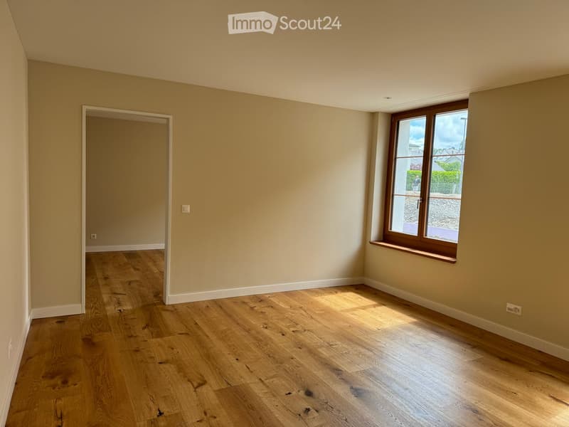Empty room with light wooden flooring, beige walls, and a large window with wooden frame letting in natural light. The room has a door leading to another space.
