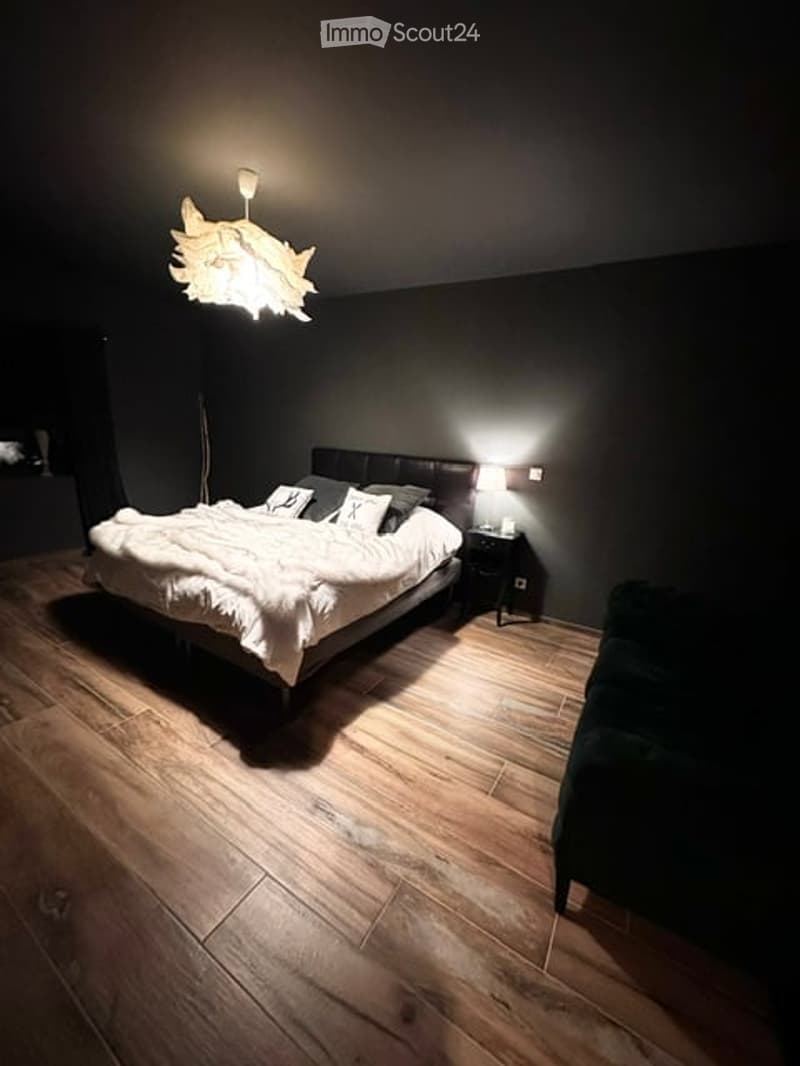 Cozy bedroom with dark walls, wooden flooring, a large bed with white bedding, a nightstand with a lamp, and a dark green armchair.