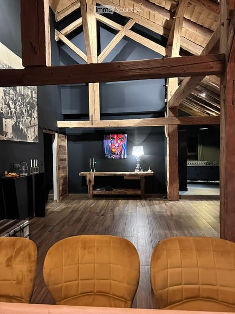A modern living room with exposed wooden beams, a large wooden table, a lamp, and a colorful abstract painting on the wall. The room features a high, wooden-beamed ceiling and warm wooden flooring.