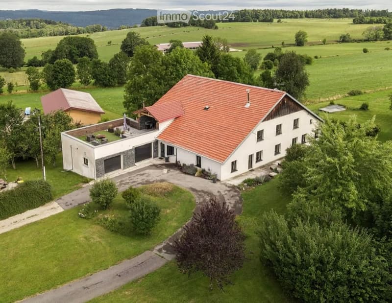 Aerial view of a two-story house with a red-tiled roof, white walls, and a large garage. The property is surrounded by green fields and trees, with a small outdoor seating area near the entrance.