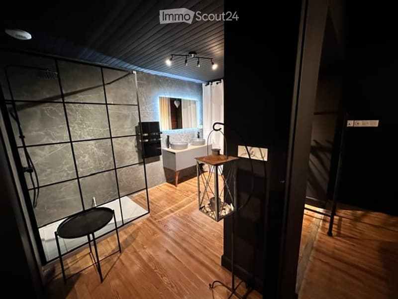 Modern bathroom with dark walls, wooden flooring, a glass-enclosed shower area, a sink with a mirror, a small table with a lamp, and a black stool.