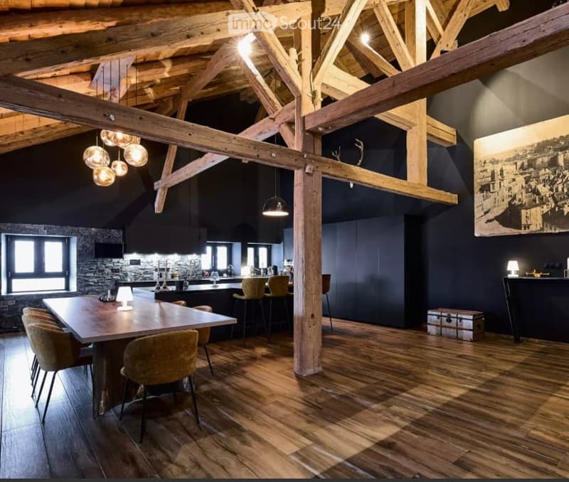Open-concept dining and kitchen area with exposed wooden beams, wooden flooring, and modern lighting. Features a large wooden dining table with chairs, a kitchen island with stools, pendant lights, and a large picture frame on the wall.