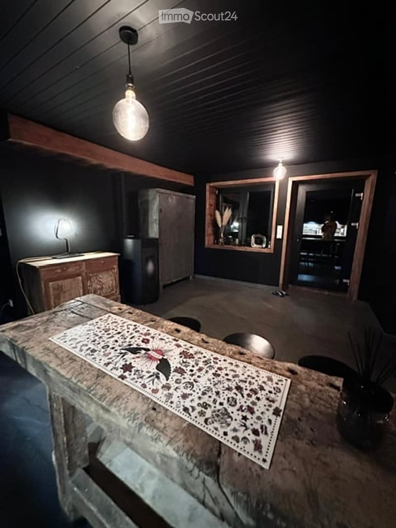 A modern, dimly lit room with a rustic wooden table, a decorative runner, a lamp, and a cabinet. The room features dark walls and a cozy, minimalist design.