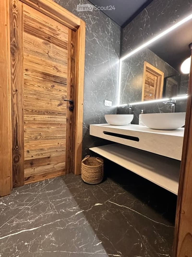 Modern bathroom with dark marble walls and floor, double sink vanity with integrated lighting, wicker basket on shelf, and wooden door.