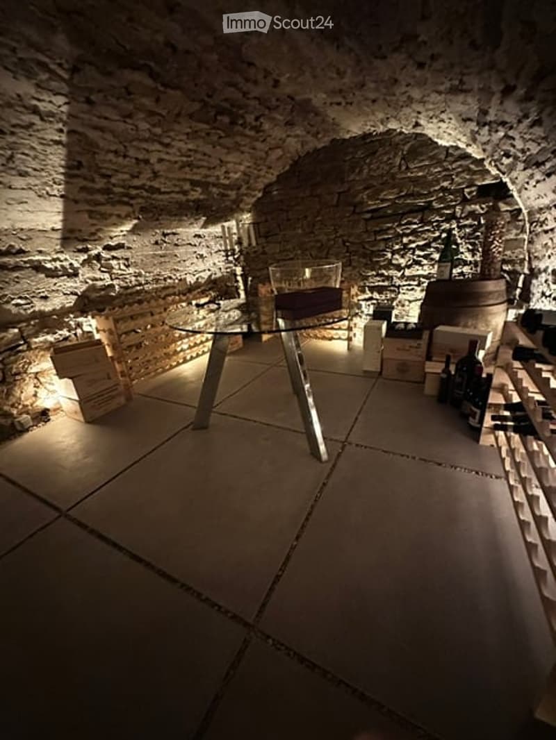 A cozy, dimly lit wine cellar with stone walls and a glass table surrounded by chairs. Wine bottles and a barrel are visible, along with a wine rack.