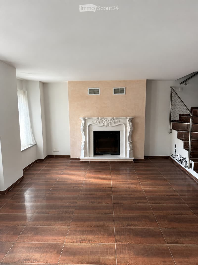 Empty room with polished wooden flooring, white walls, and a decorative fireplace. A staircase with a metal railing is visible on the right side.