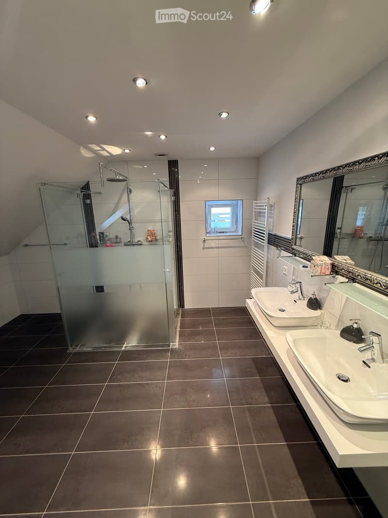 Modern bathroom with dark tiled floor, two white sinks, large mirror, glass-enclosed shower, and multiple ceiling lights.