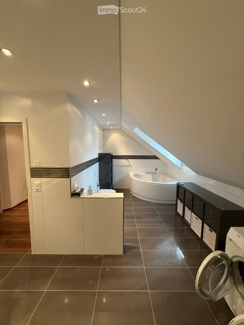 Modern bathroom with a bathtub, white cabinetry, dark tiled floor, and built-in storage. Features include recessed lighting, a skylight, and a washing machine in the corner.