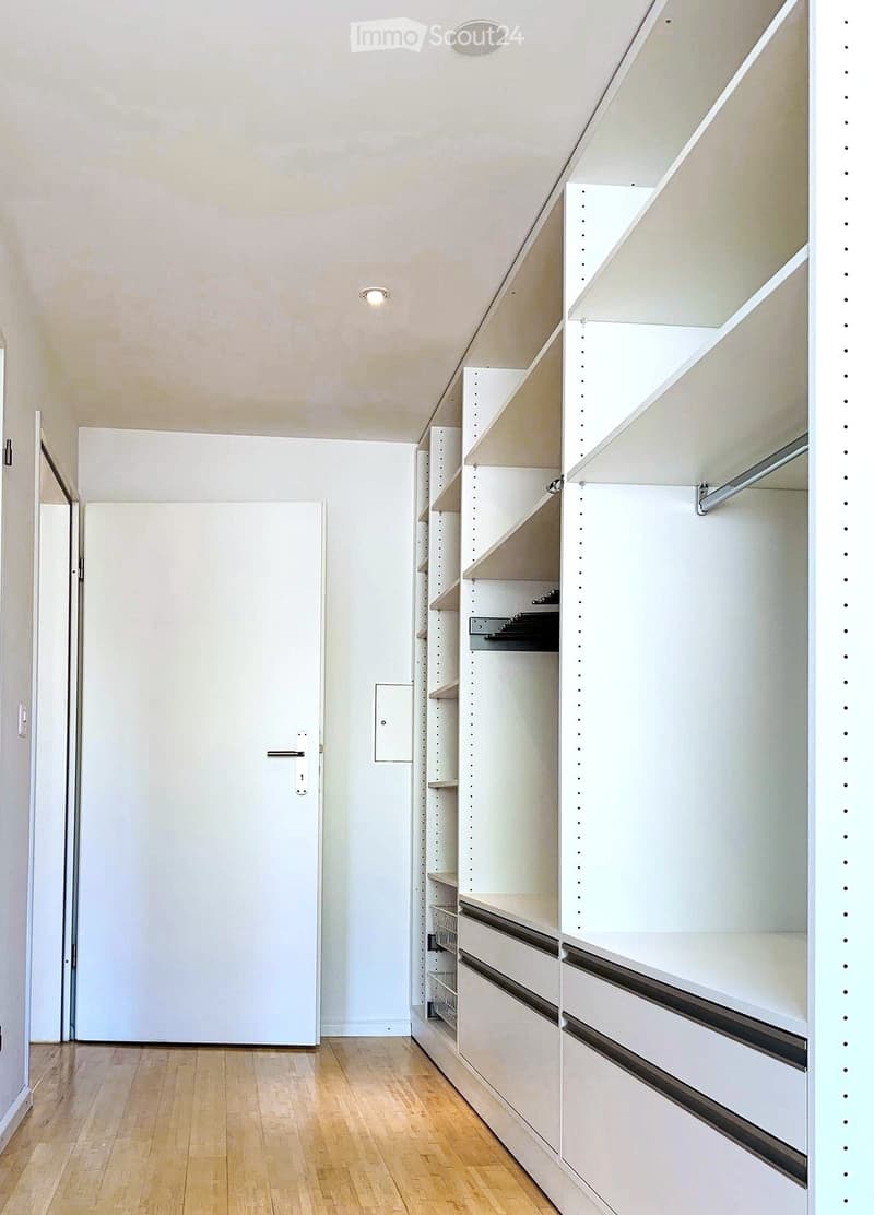 An empty, well-lit room with white walls, wooden flooring, and built-in white closet units with multiple shelves and drawers.