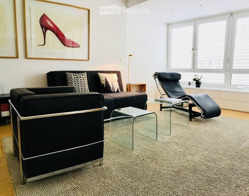 Modern living room with a black sectional sofa, glass coffee table, lounge chair, floor lamp, and artwork on the wall.