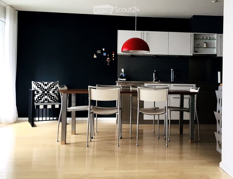 Modern dining area with a wooden table, white chairs, and a sleek kitchen area featuring white cabinets and a red pendant light.