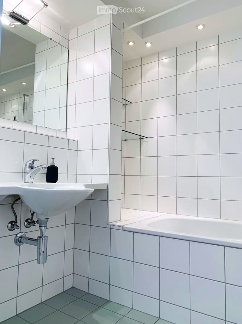 Modern bathroom with white tiled walls and floor, featuring a sink with a chrome faucet, a bathtub, and a large mirror above the sink.