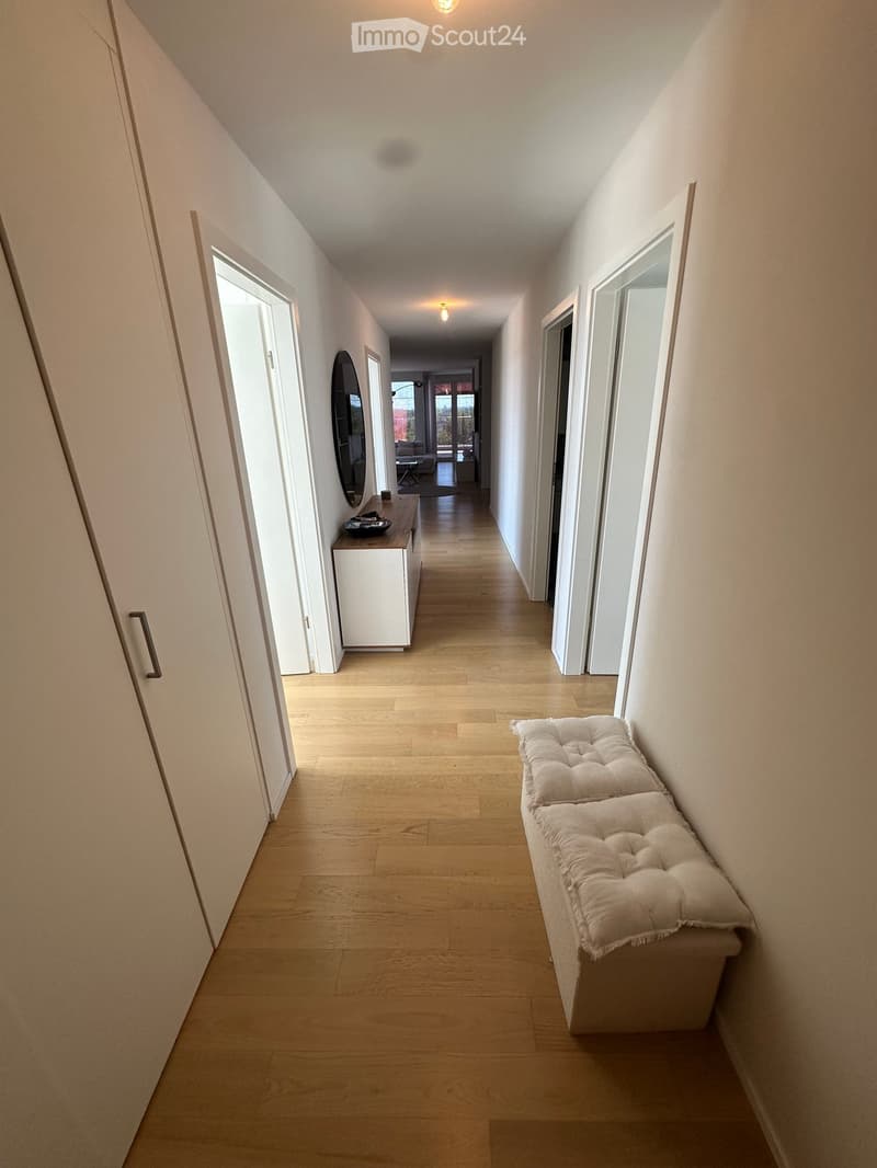 A long, narrow hallway with light wooden flooring, white walls, and recessed lighting. Features a built-in bench with a cushioned seat on one side and a small table with a mirror on the other.