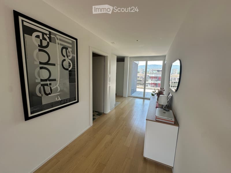 Modern hallway with wooden flooring, white walls, a large picture frame, a small console table with a lamp, and a mirror. A sliding glass door leads to a balcony with a city view.