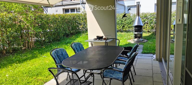 Outdoor patio with a round metal table, four blue chairs, a small table with a bowl, and a modern metal barbecue. The area is surrounded by a well-maintained lawn and hedges.
