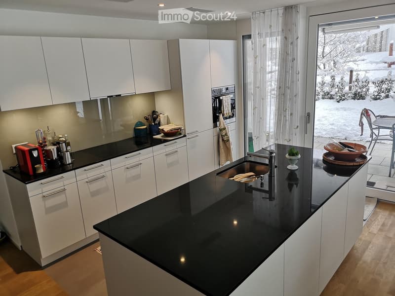 Modern kitchen with white cabinets, black countertops, and stainless steel sink. Visible items include a coffee maker, cups, bowls, and a dish rack.