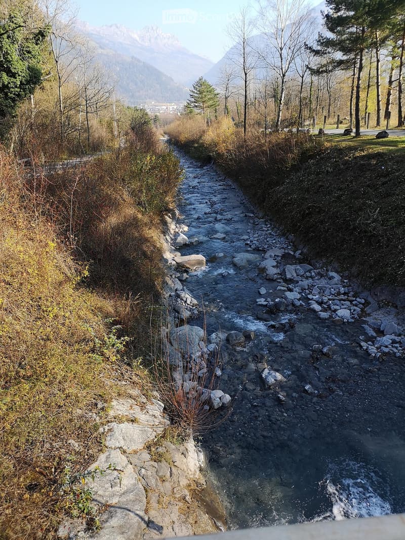A narrow stream flows through a rocky, grassy area with trees and distant mountains visible in the background.