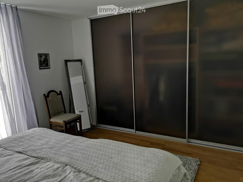 A well-lit bedroom with a large bed, a wooden chair, a standing mirror, and a picture frame on the wall. The room features wooden flooring and large sliding doors.
