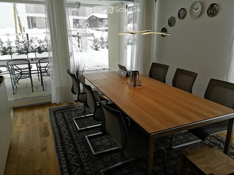 Bright room with a long wooden table surrounded by modern chairs, a laptop, and cups, with large windows showing a snowy outdoor view.