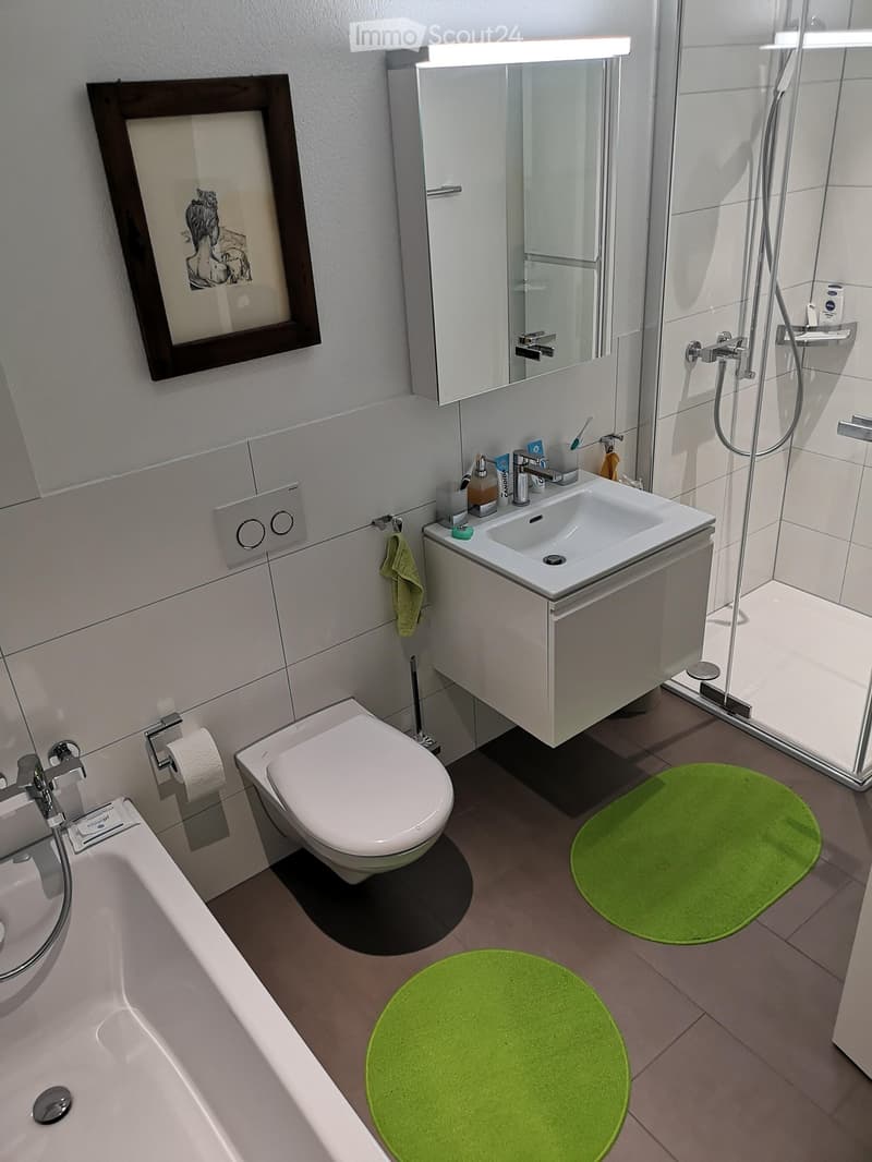Modern bathroom with white tiles, bathtub, toilet, sink, mirror, shower, and green rugs.