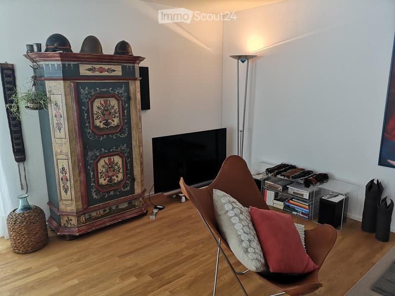 Living room with wooden flooring, a large television, a decorative fireplace, a brown leather chair with pillows, a floor lamp, and a small bookshelf with books and decorative items.
