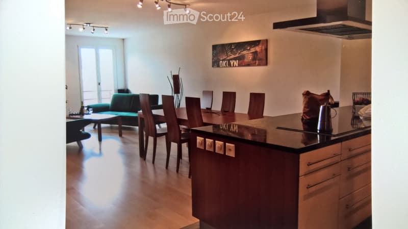 Open-plan living and dining area with a green sofa, wooden dining table with chairs, kitchen island with cabinets, and a picture frame on the wall.