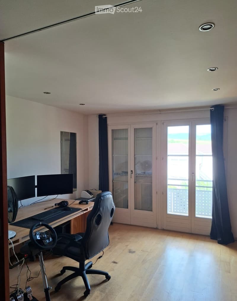 A modern, well-lit room with wooden flooring, a large desk with a computer setup, a gaming chair, and a fan. The room features large windows with curtains and a sliding glass door leading outside.