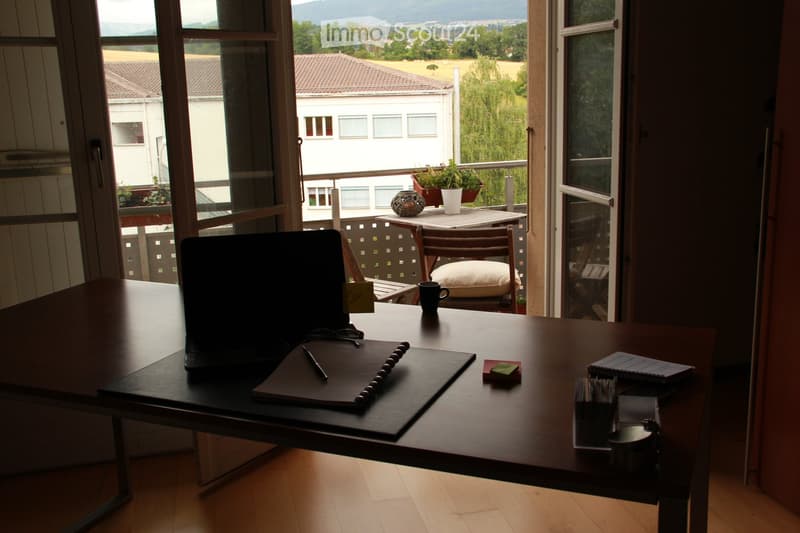 A furnished room with a wooden table, laptop, notepad, pen, cup, and a view of a balcony with outdoor furniture and a scenic landscape.