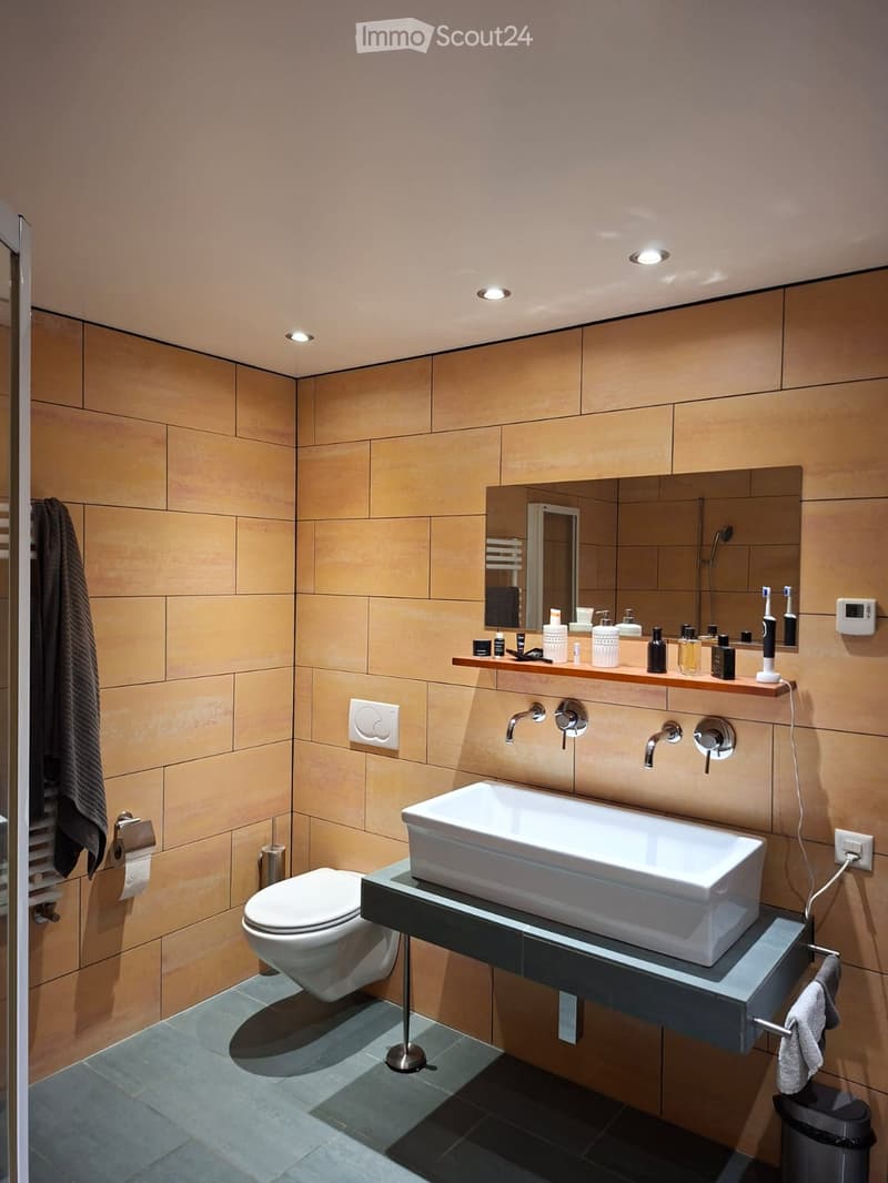 Modern bathroom with beige tiled walls, a white sink, a toilet, a large mirror, and a wooden shelf holding toiletries.
