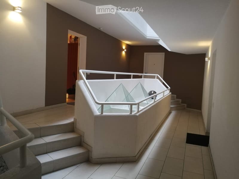 Modern hallway with tiled flooring, white railing, and a small skylight above. Features warm lighting and a door leading to another room.