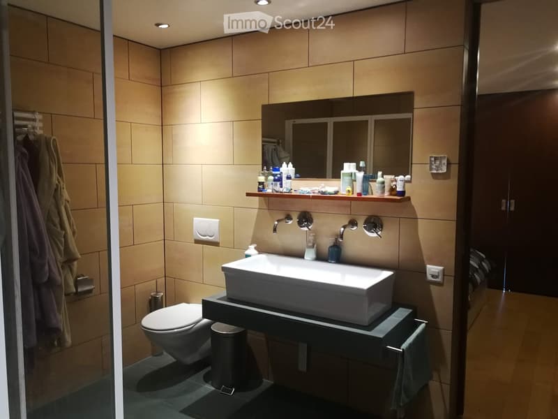Modern bathroom with beige tiles, a large sink, toilet, mirror, and various toiletries on a shelf.