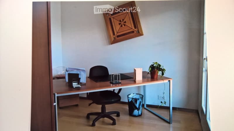 A modern, well-lit room with a wooden desk, black office chair, laptop, small potted plant, and a decorative framed artwork on the wall.