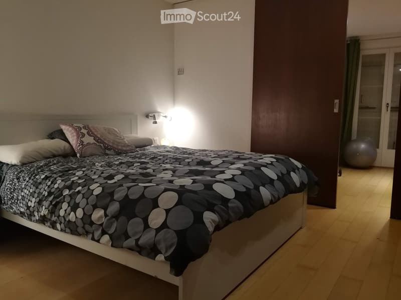 A modern bedroom with a white bed frame, patterned bedding, and a wall-mounted light fixture. A large exercise ball is visible in the adjacent space.