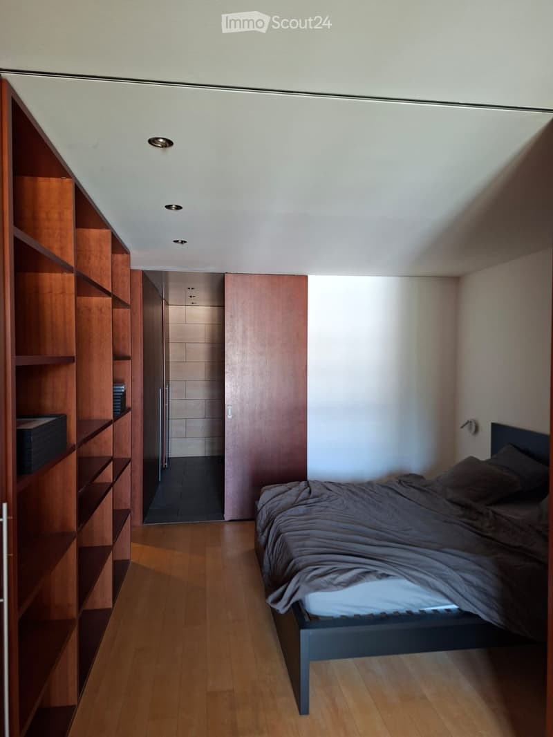 Modern, compact bedroom with a slanted ceiling, wooden floor, and built-in wooden shelving unit. Features a bed with dark bedding, a wardrobe, and a small adjacent bathroom.