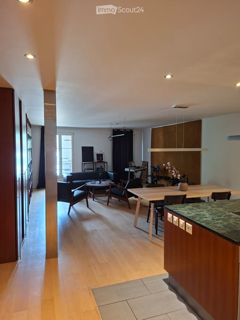 Open-plan living and dining area with wooden flooring, modern furniture including a sofa, coffee table, dining table, chairs, and a kitchen counter. A laptop and guitar are visible, along with a flower vase on the counter.