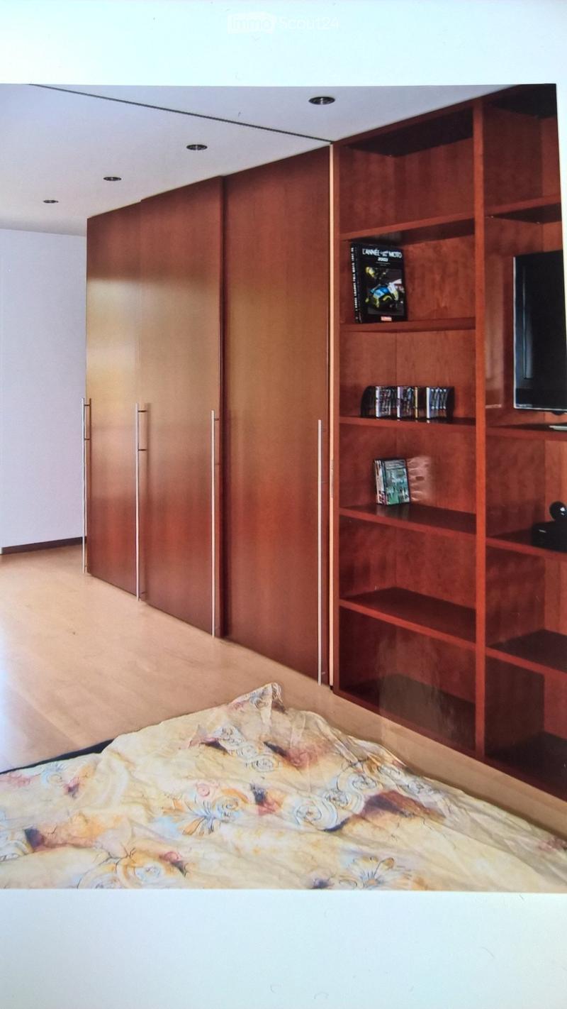 A modern bedroom with a large bed covered in a floral duvet, a tall wooden wardrobe with mirrored doors, and built-in wooden shelves holding books and a television.