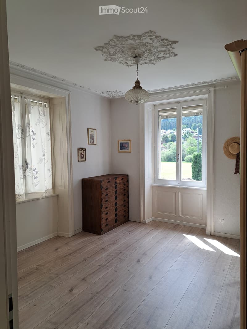 Bright room with wooden flooring, large windows, a chest of drawers, framed pictures, and a decorative chandelier.