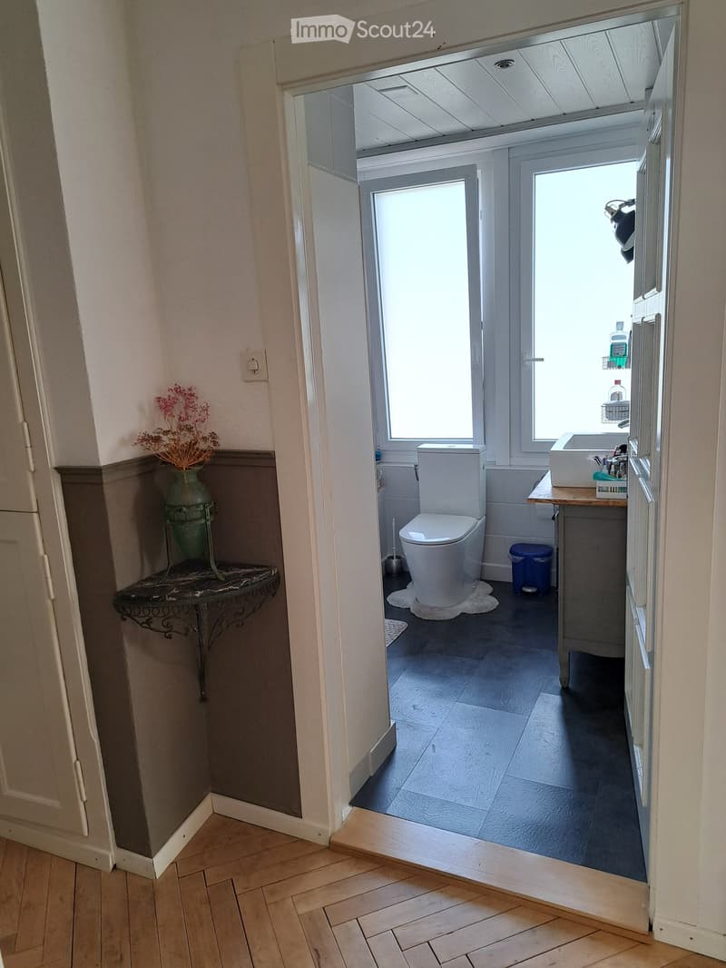 Bathroom with a toilet, sink, small table with vase, and a trash can. The room has dark tiled flooring and white walls.