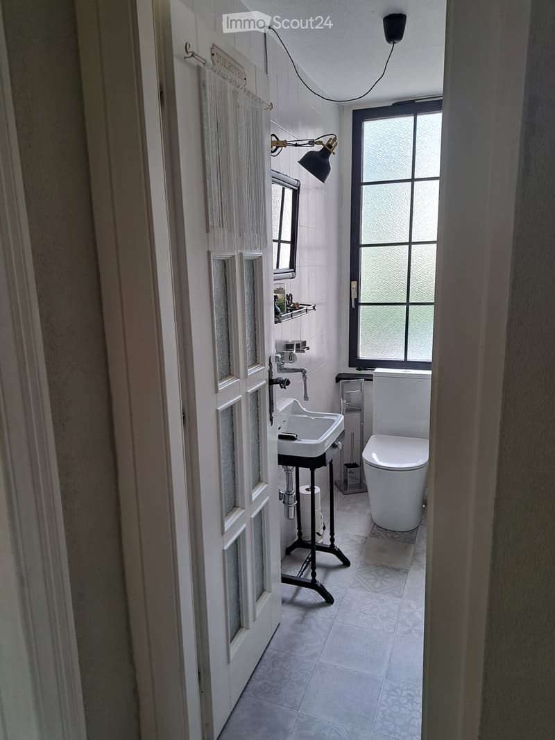 Modern bathroom with white tiles, a sink on a black stand, a toilet, a mirror, and a window with frosted glass. The room features a decorative tile floor and a wall-mounted light fixture.